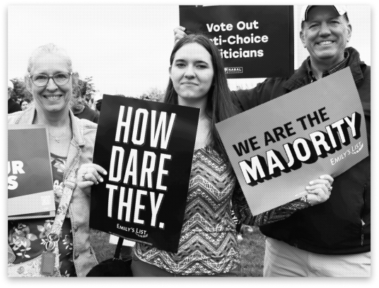 A black and white picture of three pro-choice activists, featuring rally signs that state "HOW DARE THEY" and "WE ARE THE MAJORITY" and "Vote Out Anti-Choice Politicians."